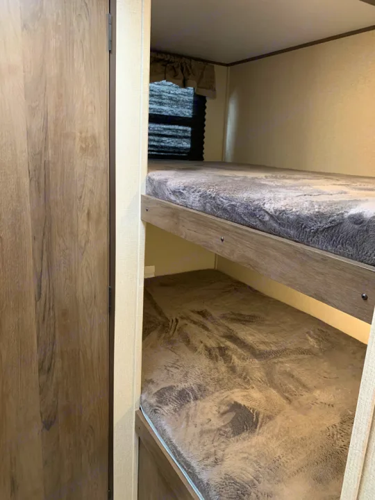RV rental bunk beds: a cozy interior showing two bunks with soft grey blankets and a window with blinds.. Prime Time Avenger 2021