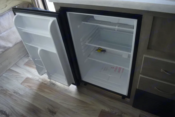 An open, clean mini-fridge inside an RV rental. Its white interior shows shelves, door storage, and some papers and keys.. Forest River X-Lite 2020