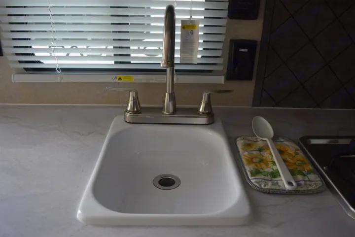 RV rental kitchen interior featuring a clean sink, faucet, window with blinds, and countertop space with a hot pad and spoon.. Forest River X-Lite 2020