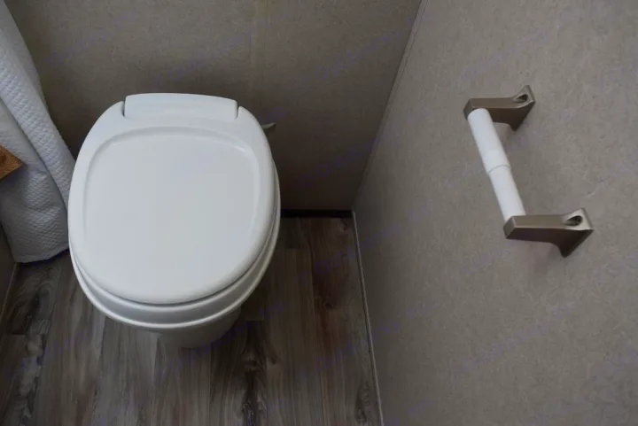 An RV rental's clean bathroom interior features a white toilet with a closed lid, wood-look floor, and a mounted toilet paper holder.. Forest River X-Lite 2020