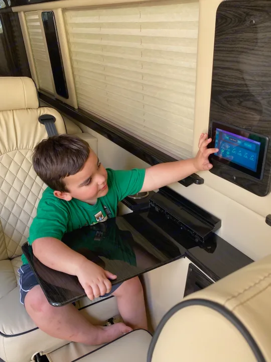 Young boy interacts with a tech control panel in a premium RV rental, featuring cream quilted seats and wood details.. Ultimate Toys Ultimate Coach 2022