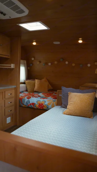 Explore a cozy RV rental interior: two inviting beds with colorful bedding, wood paneling, & ambient string lights.. Old School 818 2021