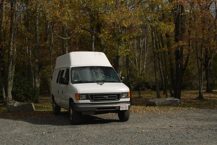 White Ford high-top van, likely an RV rental, parked on a gravel lot amidst autumn trees.. Ford E350 Box Van 2003
