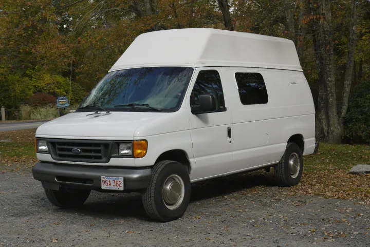 White Ford Econoline van, a high-top RV rental, parked on gravel near fall foliage.. Ford E350 Box Van 2003