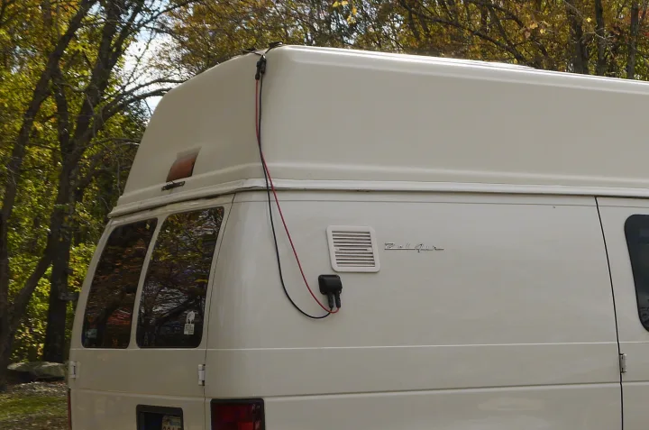 White Belaire RV rental parked amongst trees, showing its high roof, solar wiring connection, and rear windows.. Ford E350 Box Van 2003