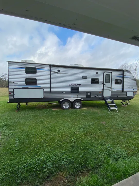 A grey & blue Catalina travel trailer, perfect for an RV rental, parked on lush green grass with steps down. Sky & trees visible.. Forest River Coachmen Catalina Destination 2019