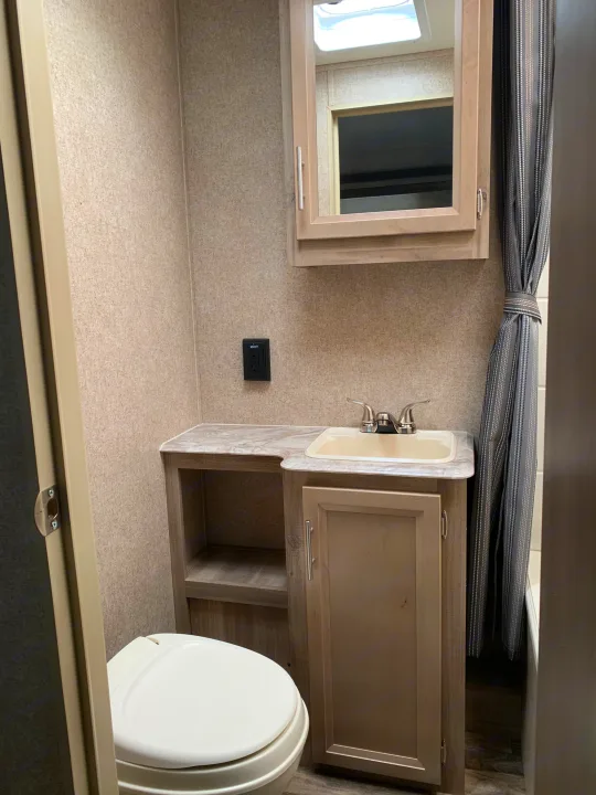 This RV rental bathroom features a compact layout with a white toilet, a sink vanity with open shelving, a mirrored cabinet, and a grey striped shower curtain.. Forest River Coachmen Catalina Destination 2019