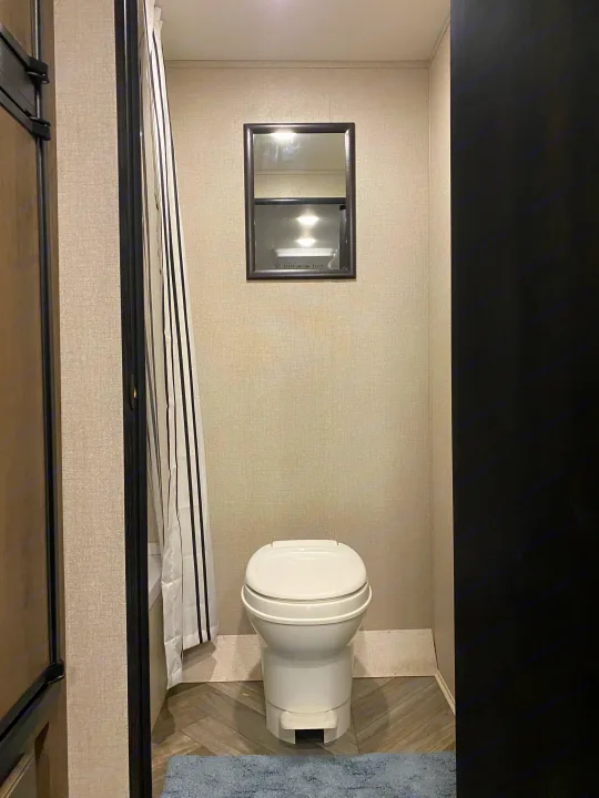 Compact RV rental bathroom showing a white toilet, mirror, striped shower curtain, and herringbone floor.. Highland ridge 19bh 2022