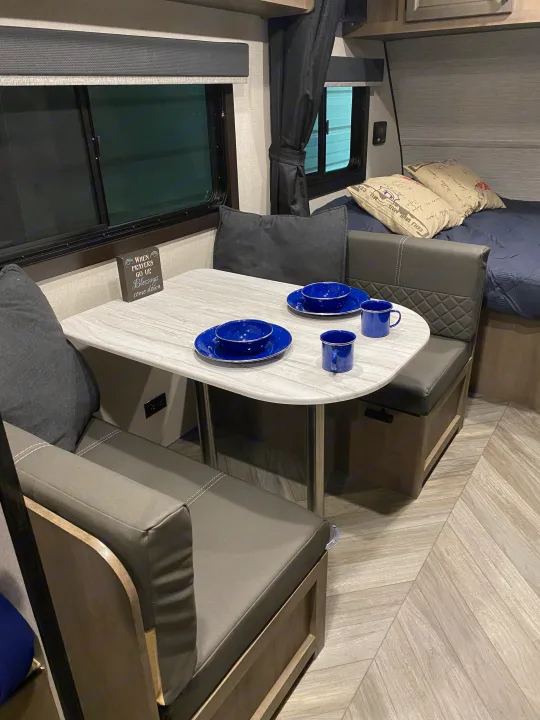 Modern RV rental interior features a compact dinette with blue dishes, gray benches, and a comfortable bed area.. Highland ridge 19bh 2022