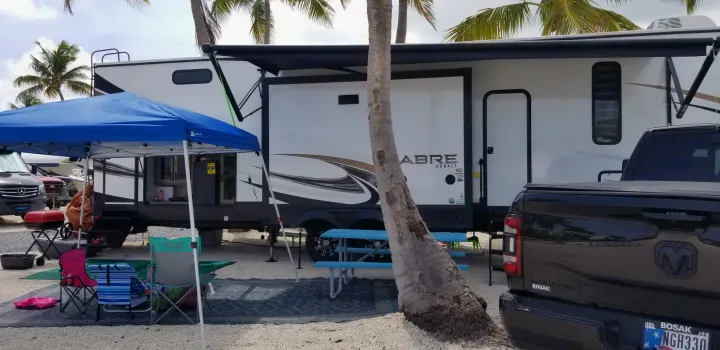 An RV rental, a white Sabre Cobalt travel trailer, parked at a gravel campsite with a blue canopy and outdoor living space.. Forest River Sabre 2021