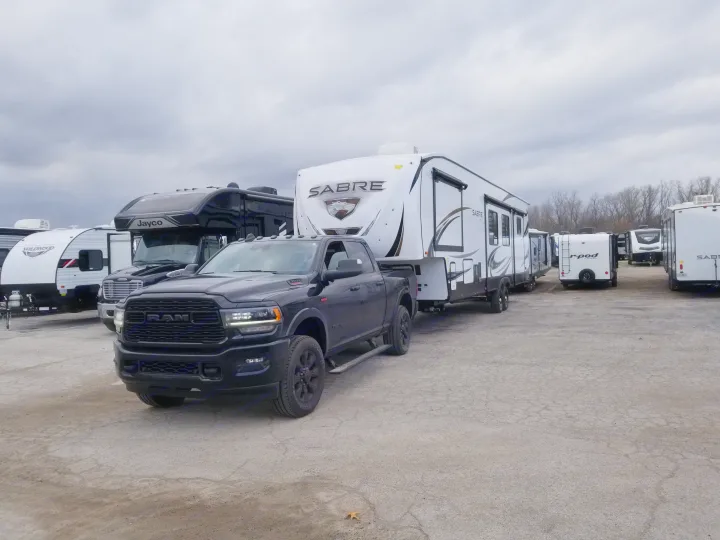 A black Ram pickup tows a Sabre fifth-wheel RV, with various RV rentals and campers parked on a lot under a cloudy sky.. Forest River Sabre 2021