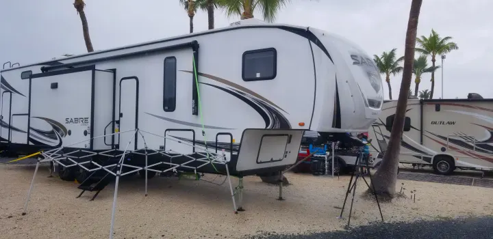A large white fifth-wheel RV (Sabre) and a motorhome (Outlaw) are parked at a gravel RV site with palm trees, perfect for an RV rental.. Forest River Sabre 2021
