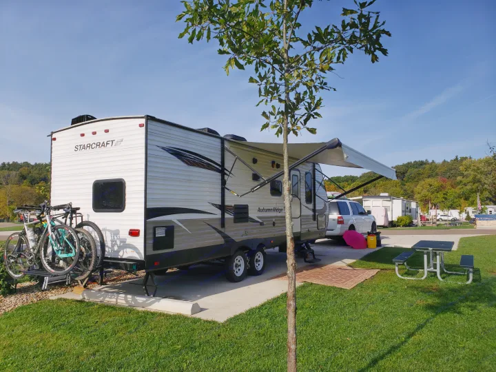 A white Starcraft rv rental and SUV parked at a sunny campsite, with bicycles, an awning, and picnic table.. Starcraft Autumn Ridge 2019