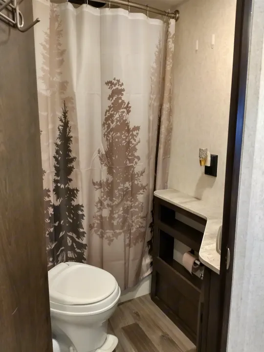 Compact RV rental bathroom with a white toilet, tree-patterned shower curtain, and small vanity.. Starcraft Autumn Ridge 2019