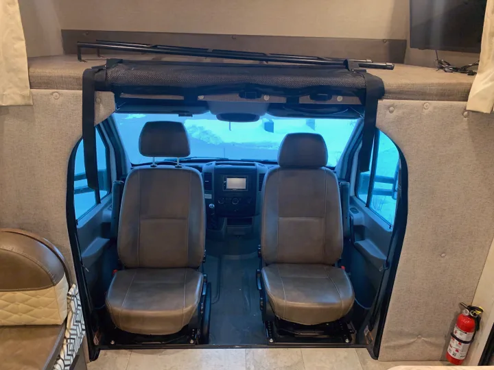 Inside an RV rental: front seats, dashboard, and an overhead sleeping area with a safety barrier. A fire extinguisher is visible.. Forest River Coachmen Prism 2020