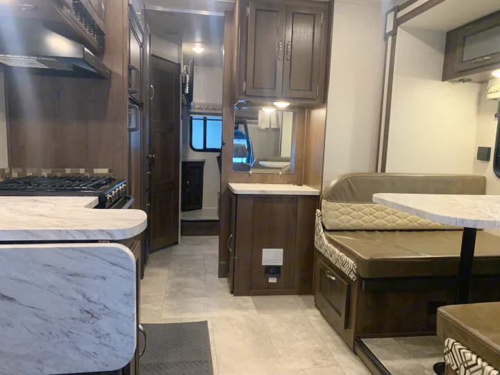 An RV rental interior: kitchen with stove, range hood, and marble counters on left. Dining booth with table on right. Hallway to rear.. Forest River Coachmen Prism 2020