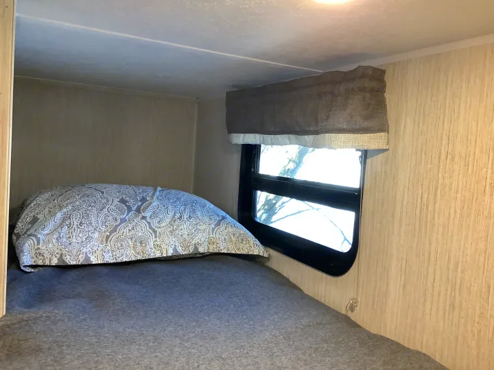 Cozy RV rental sleeping area featuring a bed with a patterned pillow, window, and a grey privacy shade.. Other Other 2021
