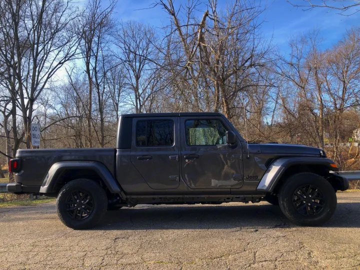 A dark gray Jeep Gladiator pickup truck, ideal for rv rental adventures, parked by bare trees on a cracked asphalt road.. Jeep Gladiator 2021