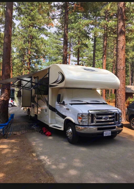 A white RV rental, a large motorhome, is parked at a shaded forest campsite with its awning extended.. Jayco Greyhawk 2016