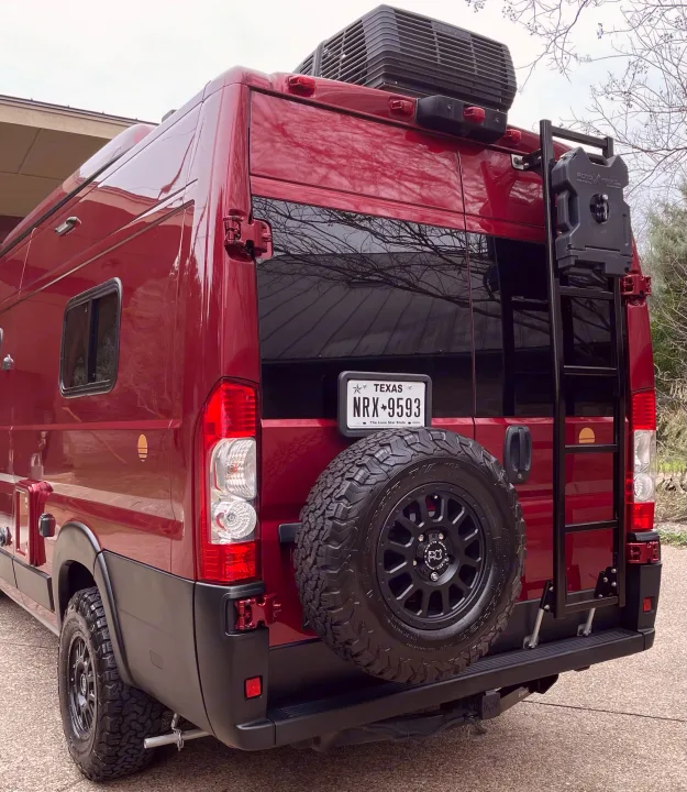 Adventure-ready red RV rental van with rugged tires, rear spare, ladder, and rooftop AC unit, shown from the back.. Winnebago Solis 2021