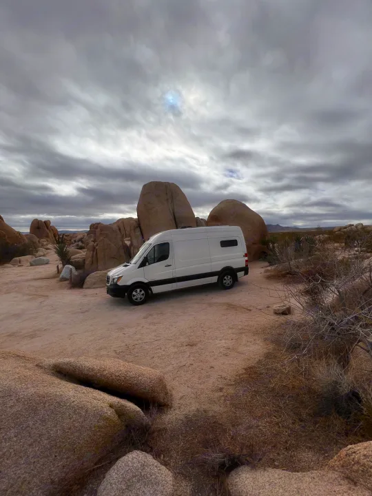 White RV rental van parked among large rocks in a desert landscape under a cloudy sky.. Mercedes Sprinter 2024