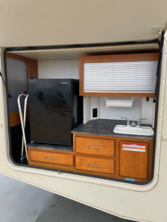Open compartment of an RV rental revealing a compact outdoor kitchen with an Insignia mini-fridge, sink, and cabinetry.. Winnebago Sightseer 2019