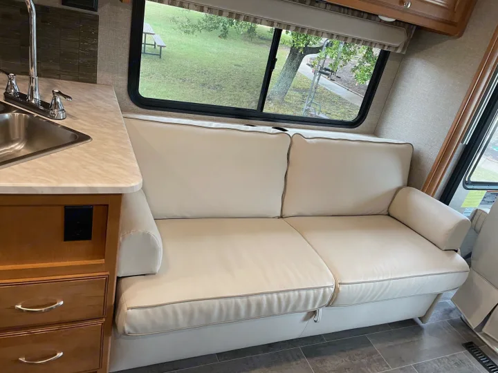 Cozy RV rental interior: cream sofa, kitchen counter with sink, and window overlooking a grassy outdoor space.. Winnebago Sightseer 2019