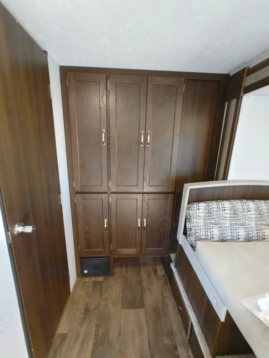 Inside an RV rental: dark wood storage cabinets, door, and a cozy bed with patterned cushion. Wood-look floor.. Keystone RV Springdale 2020