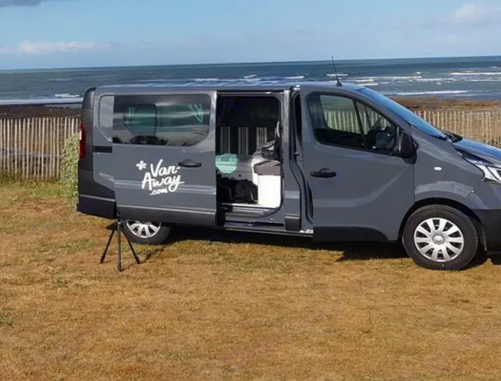 A gray "Van Away" camper van, available for rv rental, is parked seaside with its side door open, revealing the interior.. Renault Malaga 2021