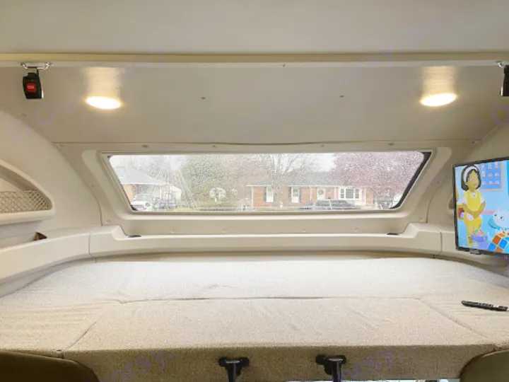 Comfy cab-over bed inside an RV motorhome rental, with a window view of a rainy street and TV entertainment.. Entegra Coach Odyssey 2022