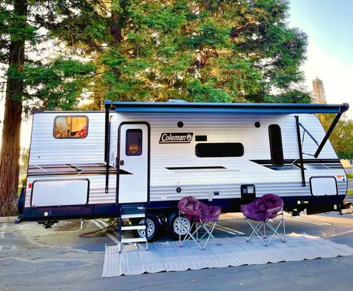 This Coleman rv rental travel trailer, with its awning extended, offers a cozy outdoor space with chairs & mat under trees.. Coleman Lantern 2022