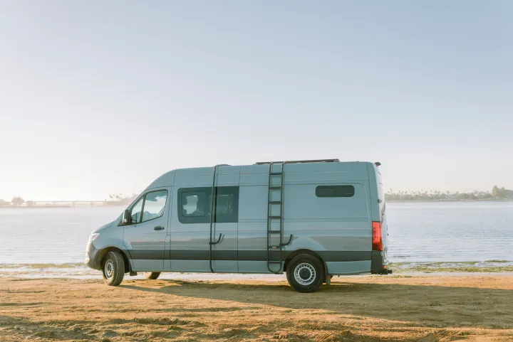 Experience freedom with this RV rental. A grey camper van overlooks a calm waterfront from a sandy beach.. Mercedes-Benz Sprinter 2021