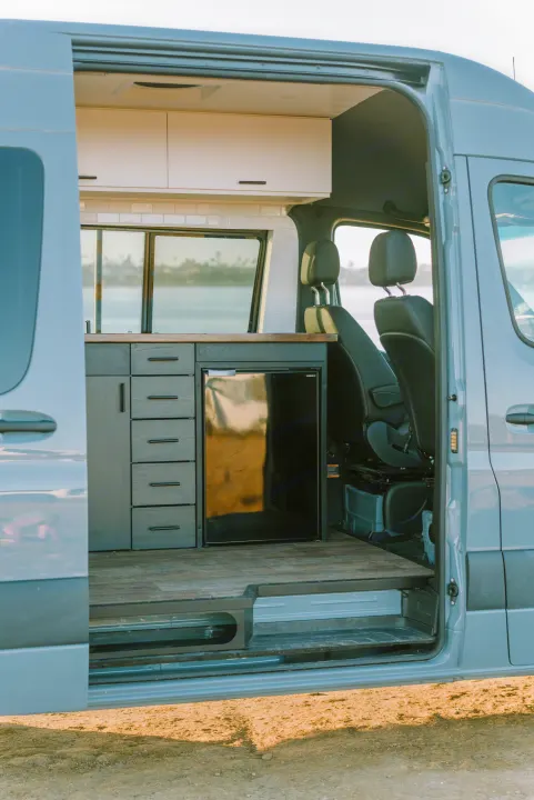 Modern rv rental interior: open side door reveals kitchen with dark cabinets, fridge, and wood floor, with water view.. Mercedes-Benz Sprinter 2021