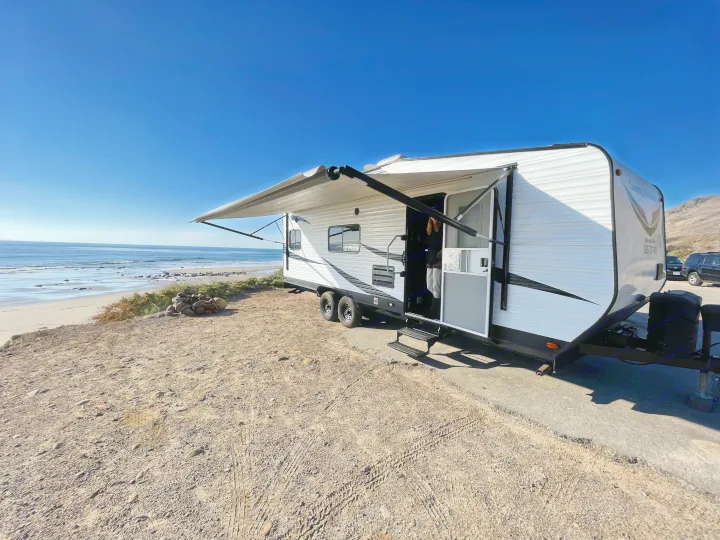 Coastal RV rental setup: Travel trailer with awning out, parked by the beach and ocean. Person visible inside.. Dune Sport Toy Hauler 2017