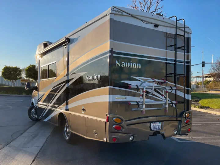 A brown and dark green Winnebago Navion RV rental, featuring a rear ladder and bike rack, is parked outdoors on a sunny day.. Itasca Navion 2016