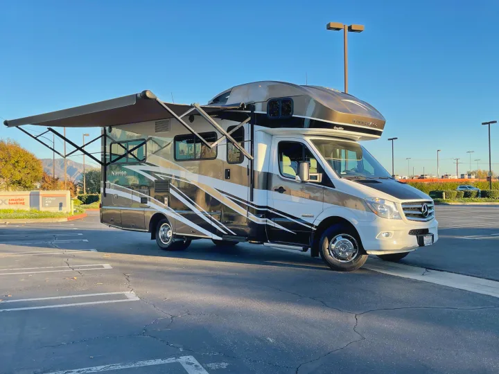 Winnebago Navion RV rental parked with awning extended in a lot under a clear blue sky, mountains visible.. Itasca Navion 2016