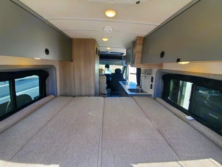 This RV rental camper van interior features a large bed, overhead storage, and a compact kitchen counter with a sink.. Winnebago Other 2022