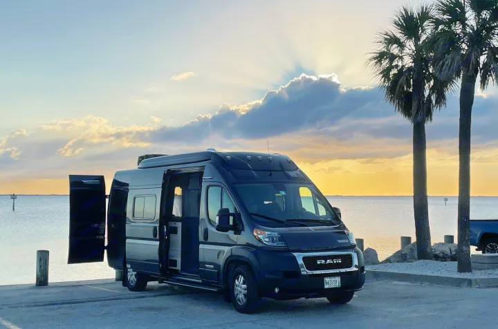 This dark RV rental camper van rests by calm water at sunset, with sun rays piercing clouds and palm trees silhouetted.. Winnebago Other 2022