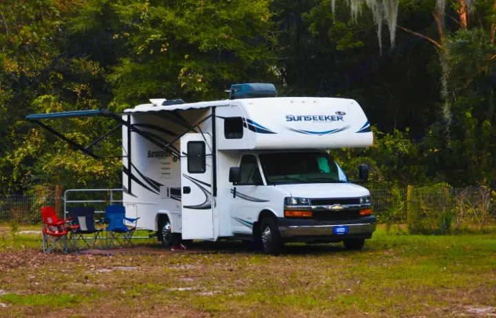 A white Sunseeker Class C motorhome, an ideal rv rental, is parked at a grassy campsite with an extended awning and outdoor chairs.. Forester LE 2018