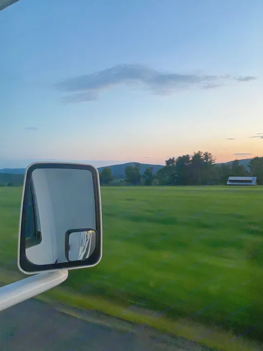 RV rental road trip at dusk: a scenic view of green fields, mountains, and sunset sky, framed by the vehicle's side mirror.. Thor Four Winds 2021