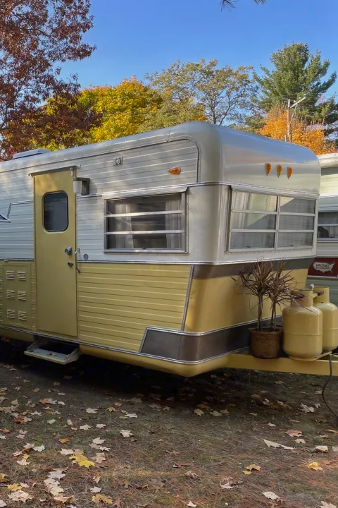 Explore this charming yellow & silver vintage RV rental. A classic travel trailer parked among autumn leaves under blue sky.. Holiday Rambler Traveler 1964