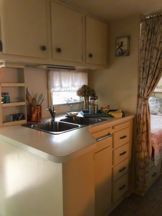 Inside a charming vintage **RV rental**: a compact kitchen with double sink, white cabinets, and a glimpsed bedroom.. Holiday Rambler Vacationer 1969