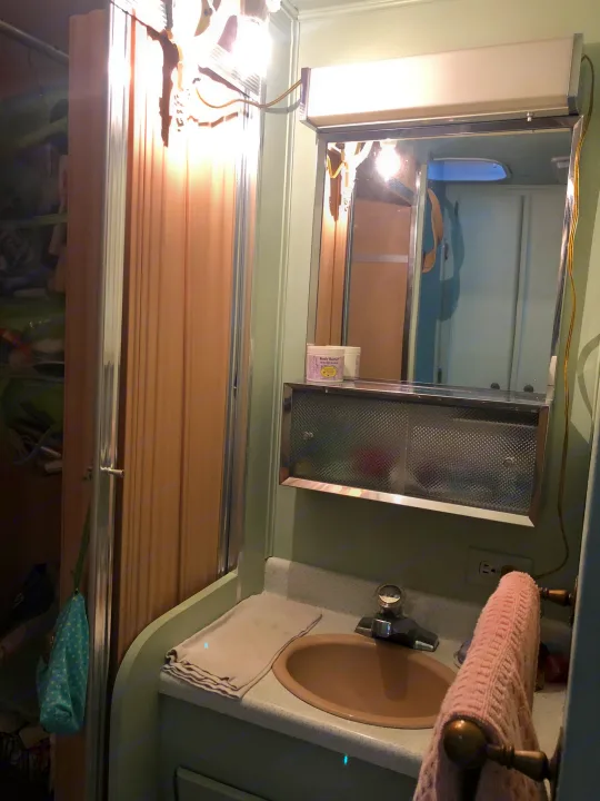 RV rental bathroom featuring a compact vanity with a beige sink, illuminated mirror, accordion shower, and storage visible on the left.. Holiday Rambler Vacationer 1969