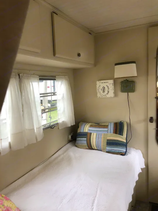 This RV rental interior features a cozy single bed, wall lamp, and window with curtains, offering a compact stay.. Holiday Rambler Vacationer 1969