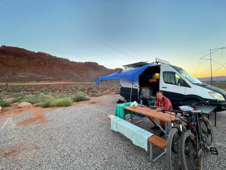 An RV rental camper van setup at a desert campsite. A person sits at a picnic table with bikes nearby.. Dodge Sprinter Van 2008