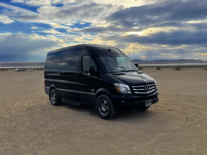 Black Mercedes-Benz Sprinter van, great for RV rental, parked on desert land. Distant lake, mountains & rainbow cloud visible.. Mercedes-Benz MB140 2015