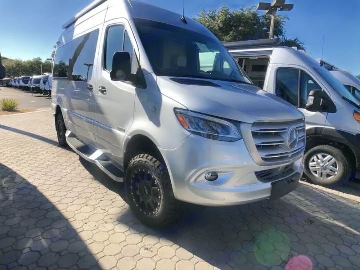 Luxury silver Mercedes-Benz Sprinter RV rental, a modified camper van with off-road tires, parked on a sunny lot.. American Coach Patriot 2022