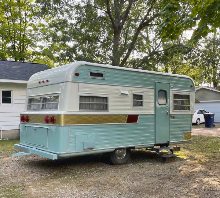 A beautifully restored vintage travel trailer, teal, white, and gold, parked on gravel. Consider this unique rv rental.. Holiday Rambler Traveler 1965