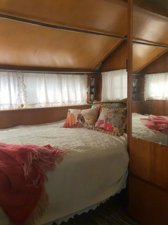Cozy RV rental bedroom features a comfy bed with decorative pillows, sheer white curtains, and warm wood interior.. Holiday Rambler Traveler 1965