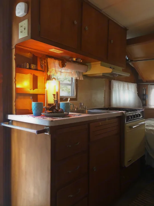 Cozy RV rental kitchen: Vintage wood cabinets, sink, stove, & inviting warm light. Perfect for camper adventures.. Holiday Rambler Traveler 1965